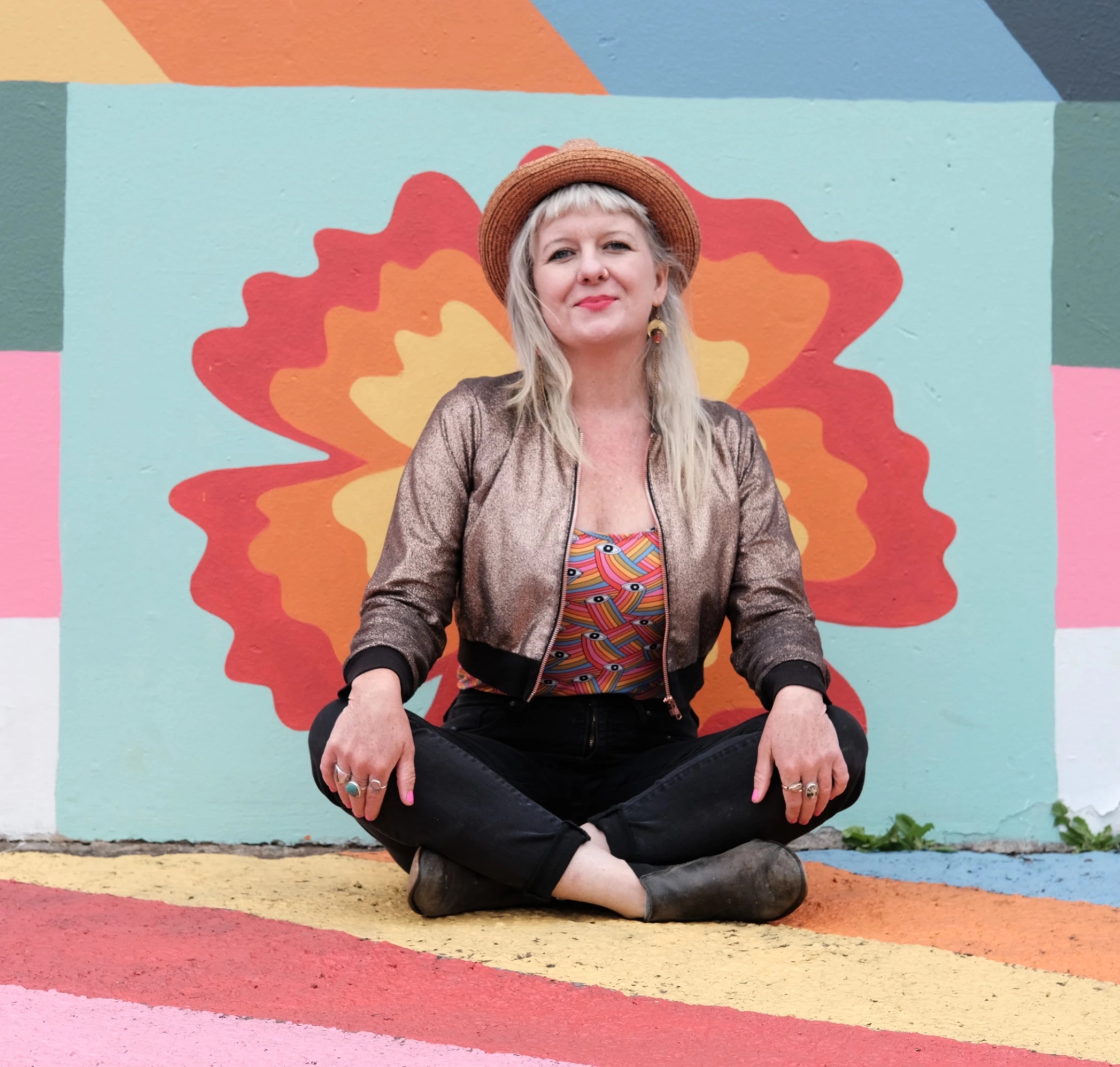 A woman with blond hair wearing a hat and shiny bronze jacket, sitting in front of a painting.