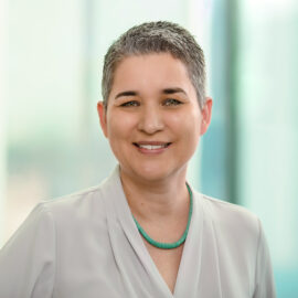 Color photograph of a woman with short, grey hair smiling, wearing an off-white blouse and a turquoise necklace