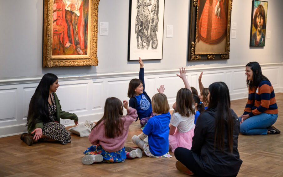 A group of children with three adults sitting on the floor of the Museum. A few children have their hands raised.