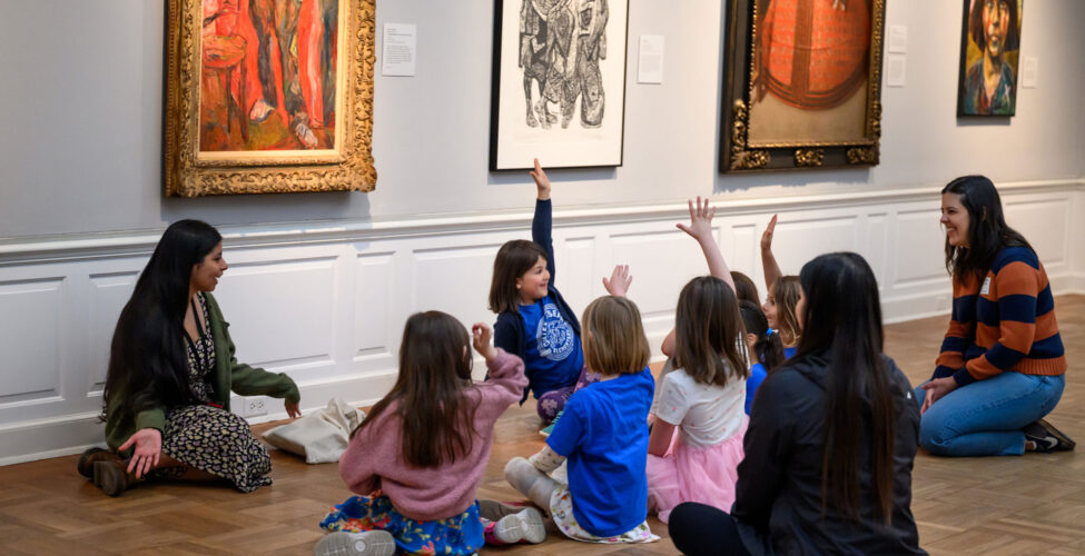 A group of children with three adults sitting on the floor of the Museum. A few children have their hands raised.