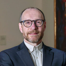 Color photograph of a man with dark eyeglasses smiling, wearing a blue jacket and white button-up shirt