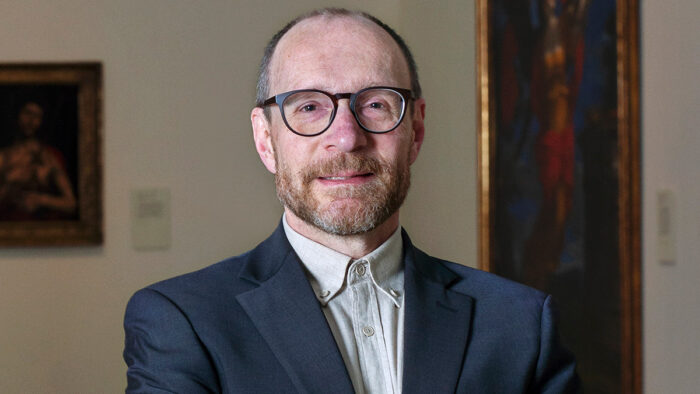 Photo of a man wearing glasses, a collared shirt and jacket, with his hands crossed in front of him.