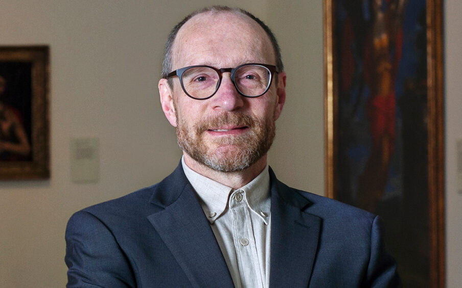 Photo of a man wearing glasses, a collared shirt and jacket, with his hands crossed in front of him.