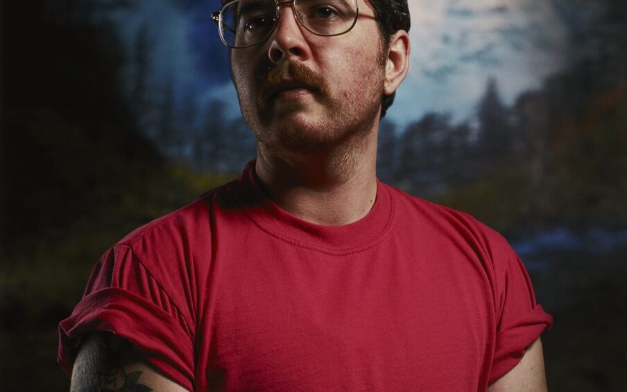 A vertical rectangular portrait of a masculine-presenting individual shown from the midsection up. The figure is positioned to face the viewer or artist and their head is turned to the left. They have light skin, dark short hair, a mustache, and beard stubble. They wear metal aviator-style eyeglasses and a red short-sleeved t-shirt with the sleeves rolled up. The figure’s bare arms show several tattoos. The upper arm at left has the words “MOM” and “DAD” within a heart design and another is of an insect. On the figure’s forearm at right, part of a bird tattoo is visible. The background is slightly out of focus and depicts large, jagged, snow-covered mountain peaks in blues and whites. Below the mountains are pine trees and dark foliage.
