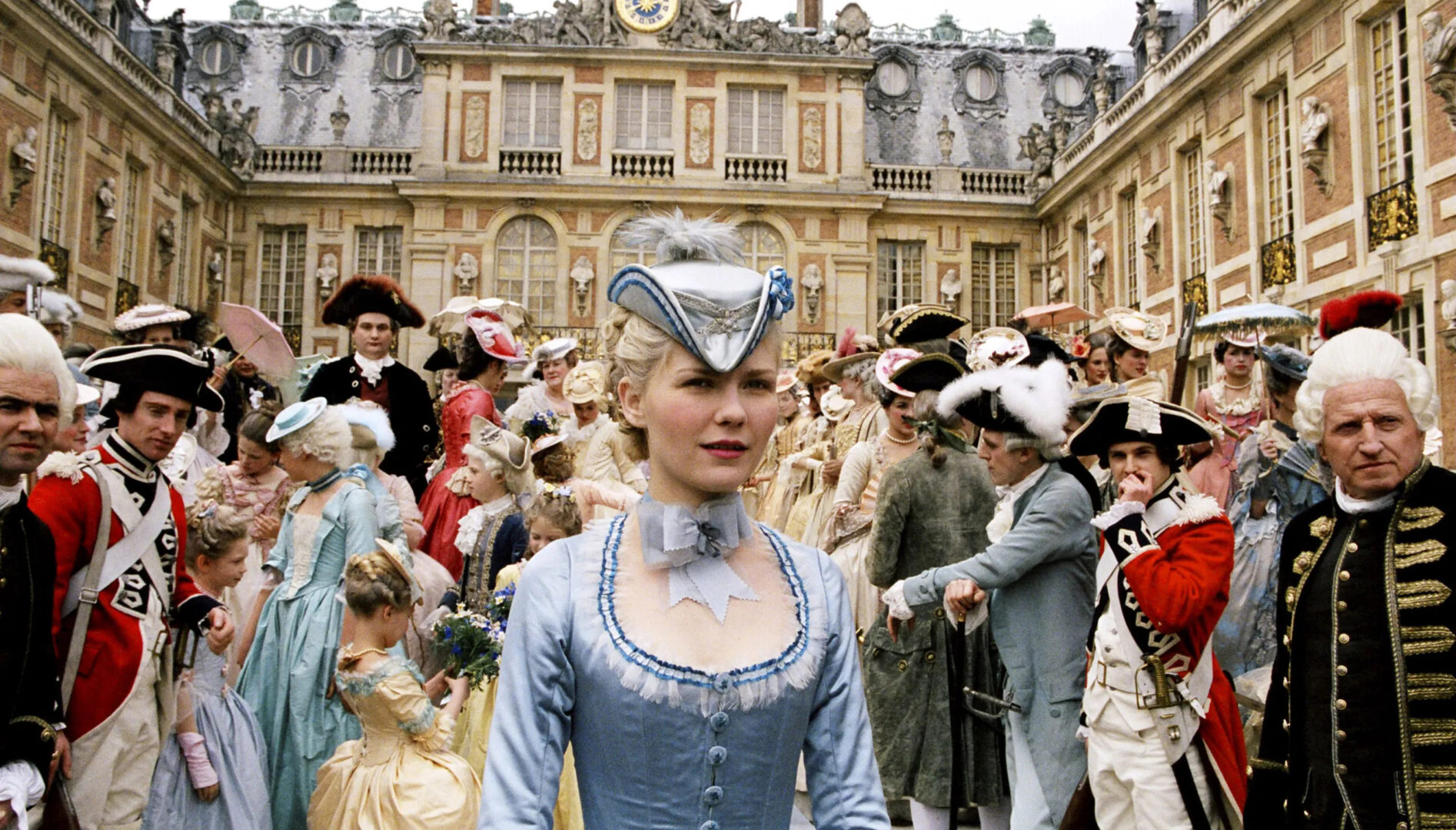 Film still of Marie Antoinette in a light blue dress, standing in front of a palace with a crowd of people behind her