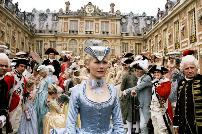 Film still of Marie Antoinette in a light blue dress, standing in front of a palace with a crowd of people behind her