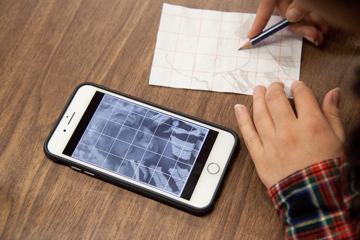 Photo of a hand drawing on lined paper next to an IPhone with an image  of a woman on it
