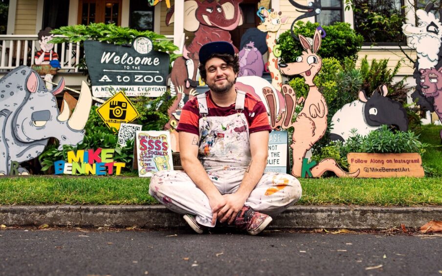 Artist Mike Bennett sitting on the curb in front of a house with his artwork on the lawn
