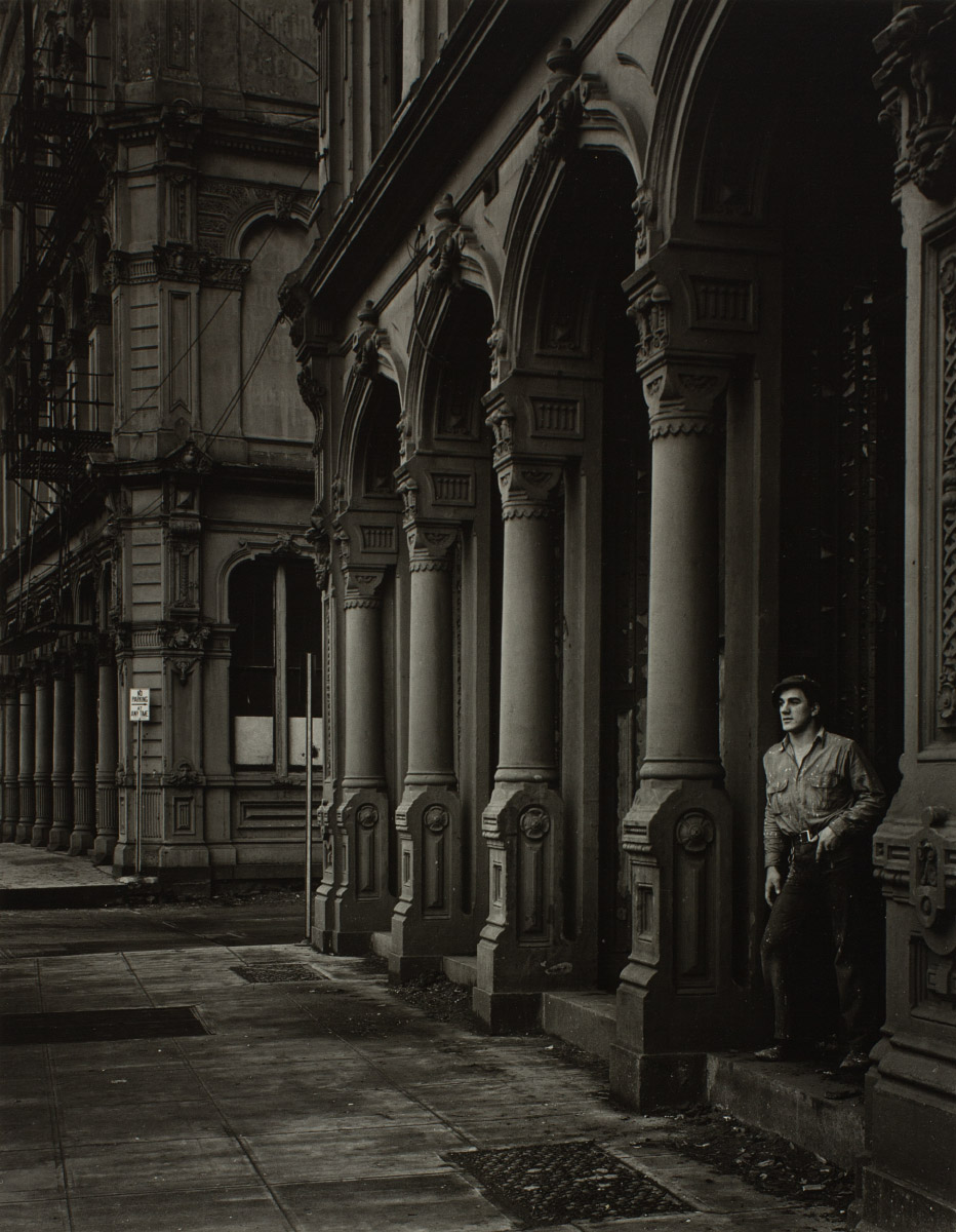 Black and white photograph of a man leaning against an arch on a city streetscape