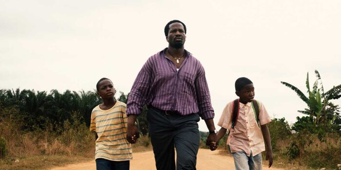 A Black man walking down a dirt road holding the hand of a young boy on each side of him.