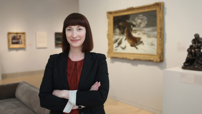 A person standing with their arms crossed in front of a painting on a wall in a gallery.