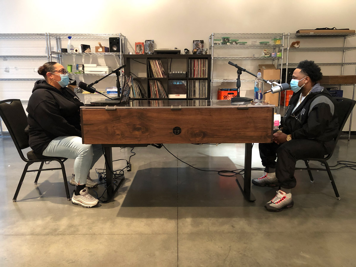A Black woman and a Black man, both wearing face masks, sitting across a wooden table from each other talking into microphones