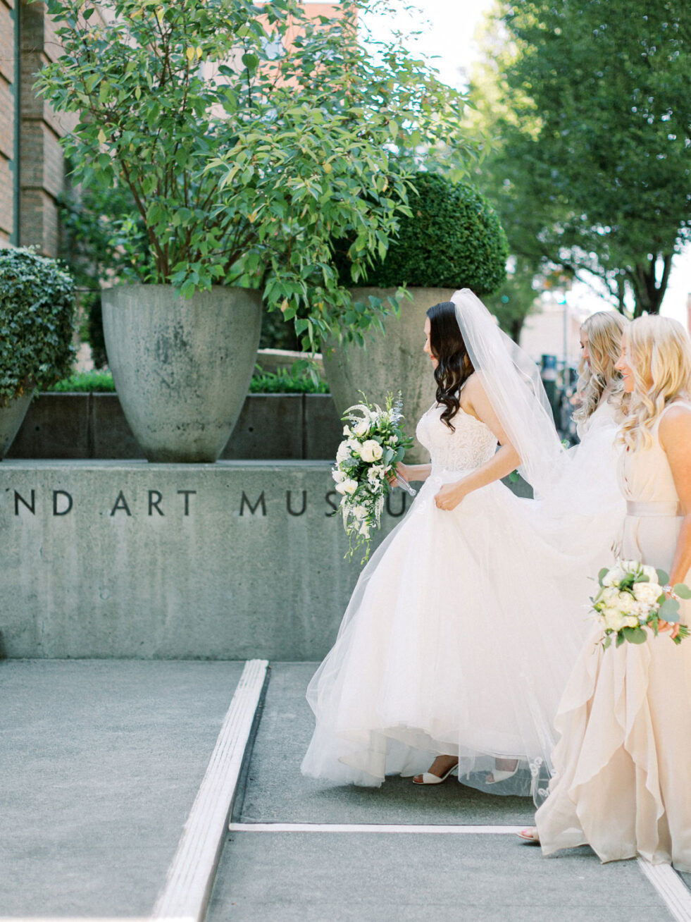 Photo of a long brown-haired bride in a white wedding dress and veil, holding a bouquet of white flowers with two women with long blonde hair and bridesmaid dresses on either side of her.