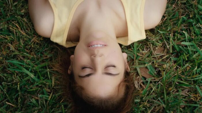 A young woman with her eyes closed and a yellow tank top on, laying on green grass.