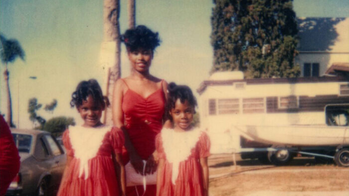 Photograph of a smiling Black woman standing behind two smiling Black girls, all wearing red dresses and standing in front of palm trees outside