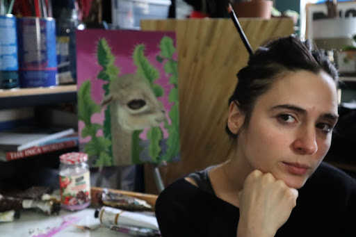 A woman with hair pulled away from her face, sitting in front of a painting resting her chin on her hand.