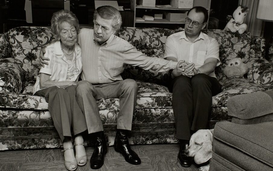 Sage Sohier, Gordon and Jim with Gordon’s Mother Margot, 1987, pigment print, 15 9/16 x 23 in. horizontal b&w photo with hot flash depicting 3 figures seated on a floral couch, the central male with his right arm around the elderly woman and left arm reaching out to male seated further away who is grasping it in both hands.