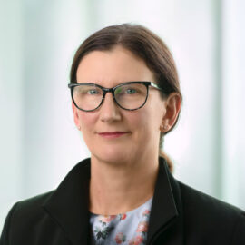 Color photograph of a woman with brown hair pulled into a pony tail, wearing eyeglasses, a floral blouse, and a black blazer