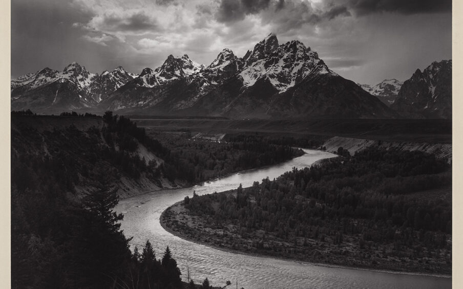 A black and white photo of a river bend with trees in the foreground and mountains in the distance