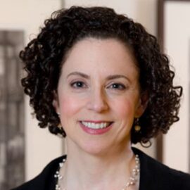Color photograph of a woman with curly brown hair smiling and wearing a pearl necklace