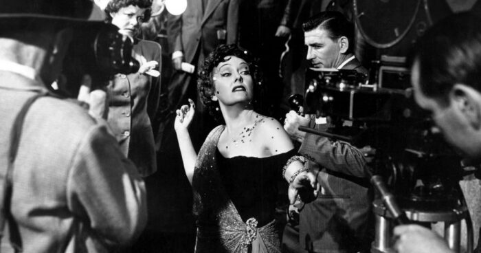Black and white film still of a woman surrounded by paparazzi.