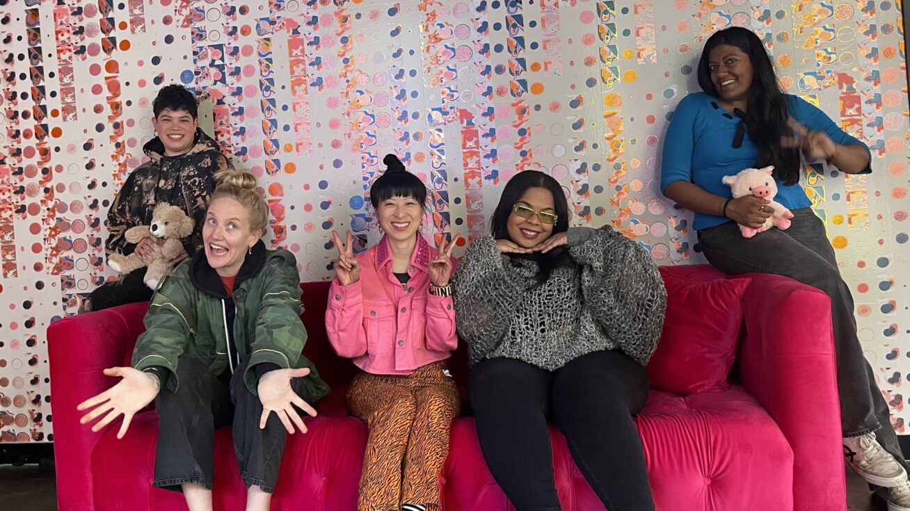 Five people sitting on and standing next to a bright pink sofa.