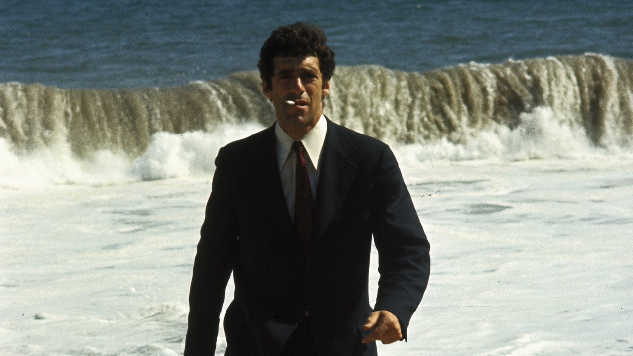A man in a suit with a cigarette in his mouth walking away from an ocean with breaking waves.