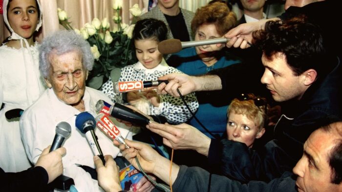 A group of reporters holding out microphones towards a very old woman.