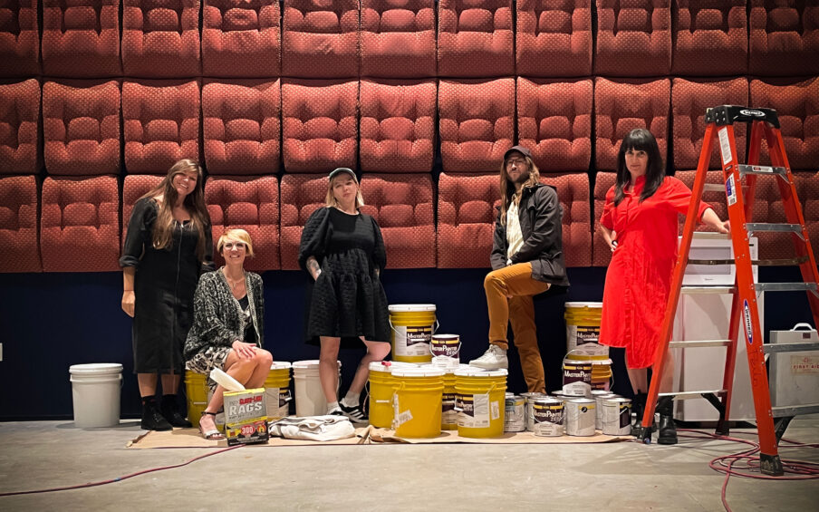 Five people standing and sitting in front of a wall covered in seat cushions. Woman at the farthest right is holding on to a red ladder.