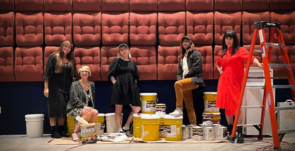 Five people standing and sitting in front of a wall covered in seat cushions. Woman at the farthest right is holding on to a red ladder.