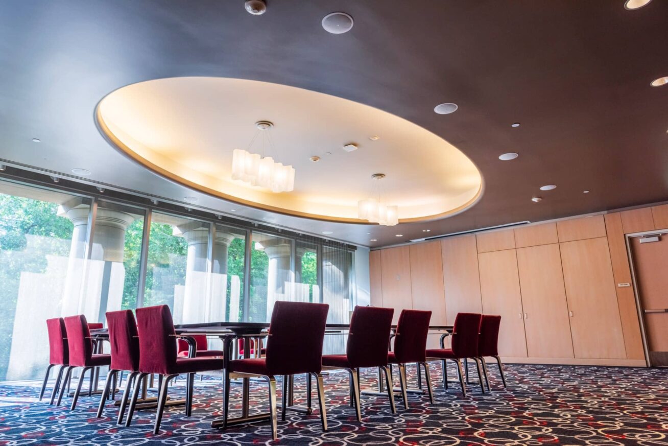A room with a large conference table surround by chairs with a wall of windows behind.