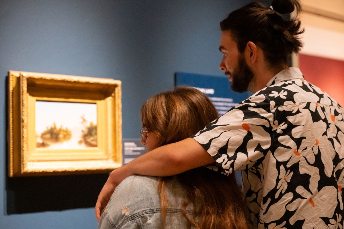 Photo of two people looking at a painting in a gallery. One has their arm around the other's shoulder.
