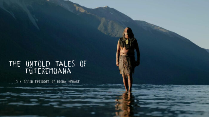 Film still of a Māori man in traditional dress, standing on a rock in the middle of water, with mountains behind him