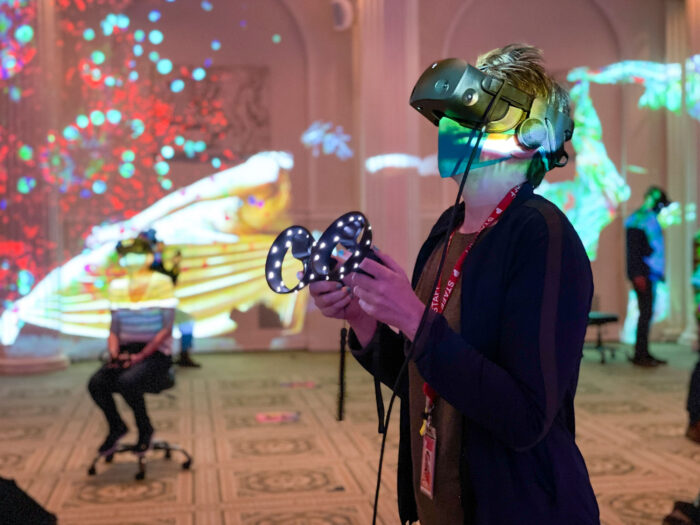 A person in the Sunken Ballroom wearing a VR headset and hand controls. Other people doing virtual reality and colored lights are in the background.