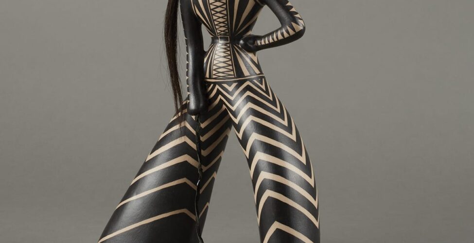 Stylized figure adorned with black-and-white triangular patterns. Details include reddish circular earrings similar to the shape of the lips, a long ponytail, and whip.