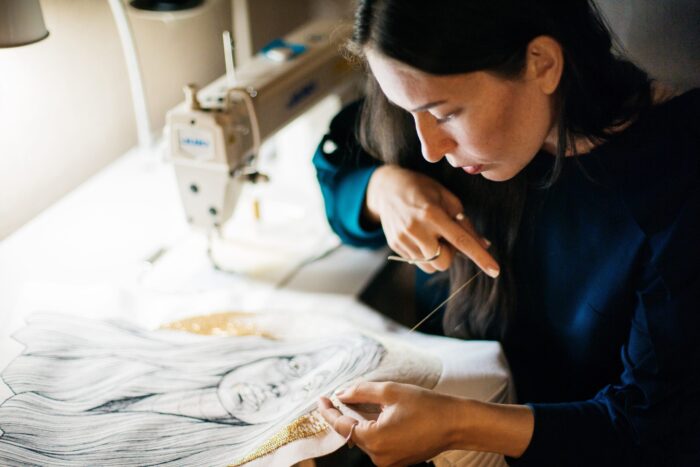 Photo of a woman with long brown hair at a sewing machine, leaning over a piece of fabric
