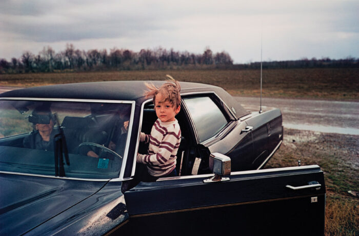 Photograph of a boy hanging out the driver's side door of a parked car. An older woman sits in the passenger's seat.