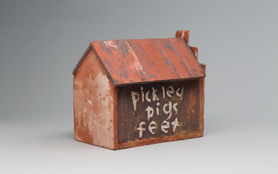 Image descriptions: Little’s Grocery (Shack #4), Willie Little, glazed ceramic, 9 ¾ x 7 ¾ x 10 ¾. 1. A three quarter view of a ceramic house with pentagon shaped ends, long sides and a sloped roof. The terracotta-colored roof has vertical ridges with deep gray and light beige portions creating a worn look. The broad side of the house is a dark brown with lighter shades of terracotta showing through. The words “pickled pigs feet” is painted on one broad side and “penny candy” on the other. The lettering is done in a lowercase, free hand style in a crackled, uneven, near white glaze with bits of terracotta showing through. The end of the house at right has a stairstep pattern rising from the gable, ending in a square shape at the peak. The short end of the house features lighter and darker terracotta colors that create texture and pattern.