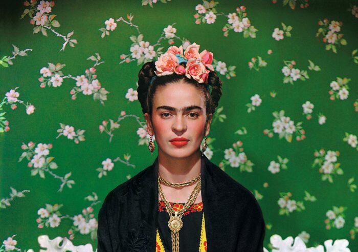 Frida Kahlo with a pink flower crown, heavy gold jewelry, and her hair up in braids, sitting against a bright green wallpaper with pink flowers on it.