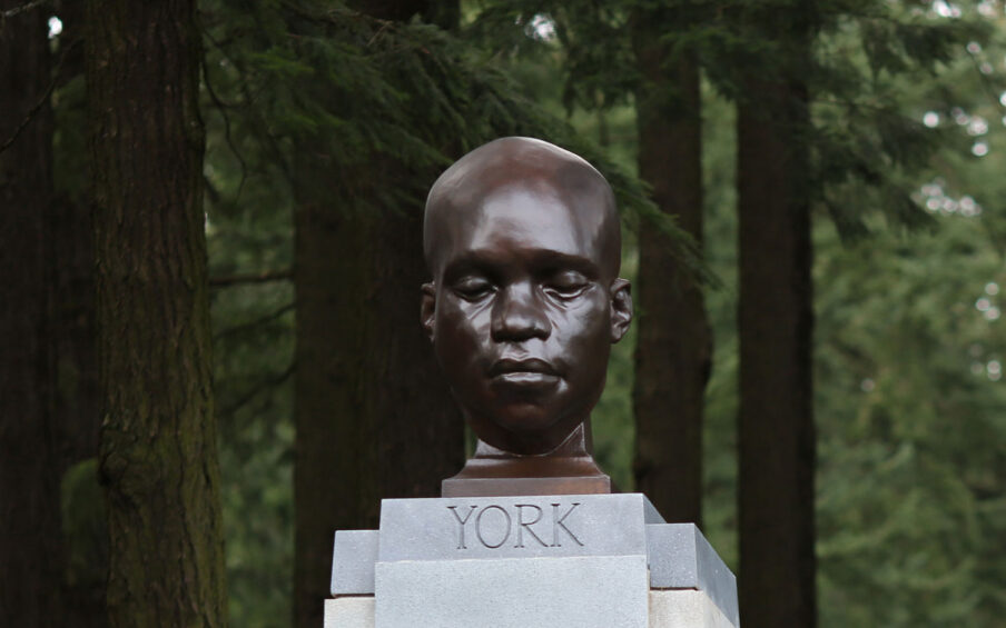 A bronze-looking head statue of a Black man with his eyes closed. It's on a stone pedestal and surrounded by big trees.