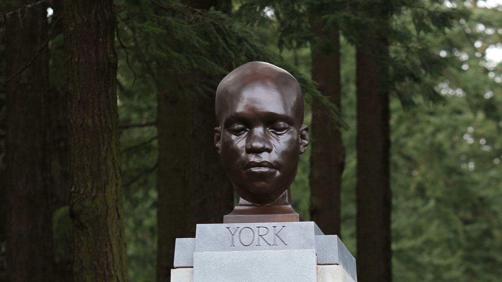A bronze-looking head statue of a Black man with his eyes closed. It's on a stone pedestal and surrounded by big trees.